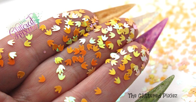 May include: A close-up of iridescent orange and yellow flower-shaped glitter. The glitter is scattered on a white surface and is being held in a hand.