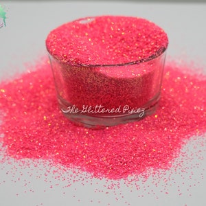 Può includere: Primo piano di glitter rosa, con una finitura fine e iridescente. Il glitter è in un barattolo di vetro trasparente, con il testo "The Glittered Pixiez" sul fondo del barattolo.