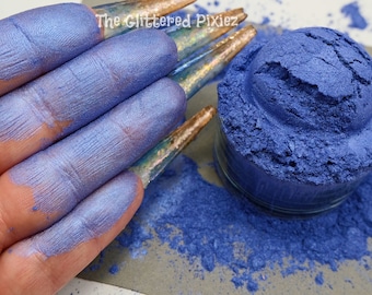Puede incluir: Primer plano de una mano cubierta de pigmento azul junto a un pequeño tarro del mismo pigmento. El pigmento es un azul metálico vibrante. El tarro transparente está sobre una superficie gris, con algo de pigmento derramado. El texto "The Glittered Pixiez" es visible.