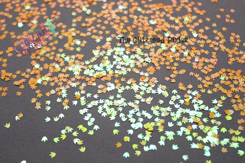 May include: A scattering of iridescent, holographic dinosaur footprint shaped glitter in shades of orange, yellow, and green. The glitter is on a dark gray background. The Glittered Pixiez is written in white text on the image.