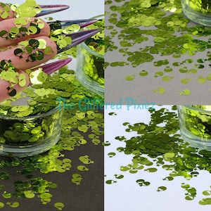 Puede incluir: Purpurina verde con forma de globo. La purpurina está en un pequeño frasco de vidrio y esparcida sobre una superficie blanca.