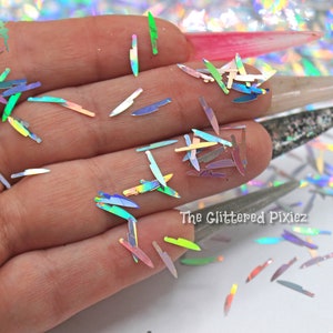 May include: A hand holding iridescent holographic glitter shaped like knives. The glitter is scattered on a white surface. The text "The Glittered Pixiez" is visible in the lower right corner of the image.