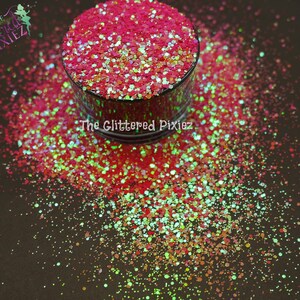 Puede incluir: Primer plano de un tarro de purpurina roja, verde y dorada. La purpurina está esparcida alrededor del tarro sobre una superficie oscura. El tarro tiene una etiqueta que dice "The Glittered Pixiez".