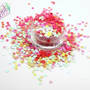 Puede incluir: Un pequeño frasco lleno de purpurina iridiscente en forma de flor en tonos de rosa, naranja, amarillo y blanco. La purpurina está esparcida alrededor del frasco sobre una superficie blanca.