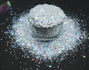 May include: A clear glass container filled with white glitter, with iridescent and silver flecks. The glitter is scattered around the container on a black surface. The text "The Glittered Pixiez" is visible in the image.