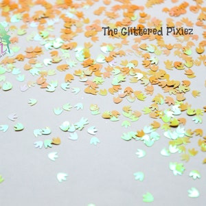 May include: A scattering of iridescent, pastel-colored, flower-shaped glitter. The glitter is a mix of light green, light blue, and light orange.
