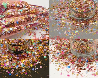 May include: A close-up of a jar of colorful glitter with a mix of heart shapes, stars, and other shapes. The glitter is scattered around the jar on a white surface.