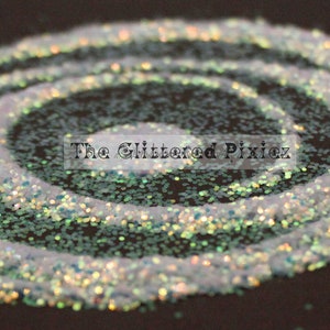 Könnte beinhalten: Ein weißer, irisierender Glitter mit einem grünen Schimmer in einem kleinen, durchsichtigen Glas. Der Glitter ist spiralförmig auf einer schwarzen Oberfläche verteilt. Der Text "The Glittered Pixie" ist auf dem Bild zu sehen.