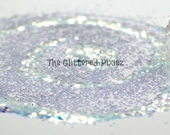 Puede incluir: Primer plano de un frasco de purpurina iridiscente con un esquema de color blanco y azul. La purpurina está esparcida alrededor del frasco sobre una superficie blanca. El texto "The Glittered Pixiez" es visible en la imagen.