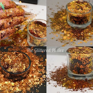 May include: A close-up of a jar filled with gold and copper glitter with a circle shape. The glitter is scattered around the jar on a white surface.