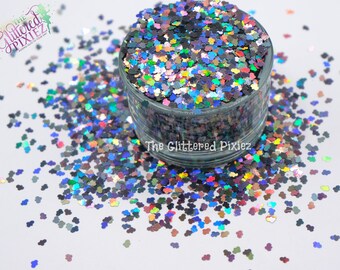 Pode incluir: Um close-up de um frasco cheio de glitter holográfico iridescente em forma de cabeças do Mickey Mouse. O glitter está espalhado sobre uma superfície branca.