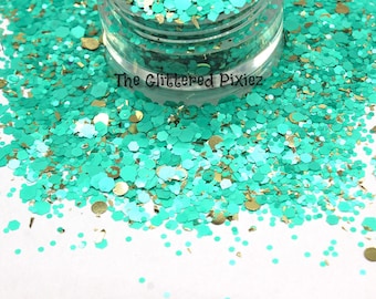 May include: A close-up of a jar of teal and gold glitter. The glitter is a mix of different sizes and shapes, including hexagons, circles, and stars. The jar is labeled "The Glittered Pixiez".