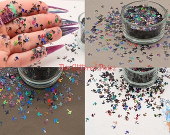 Pode incluir: Um close-up de glitter iridescente arco-íris em forma de guarda-chuvas. O glitter está espalhado em uma superfície branca e em um pequeno frasco de vidro.