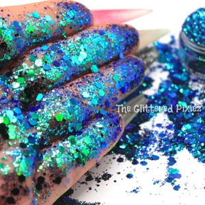 May include: A close-up of a jar of blue and green iridescent glitter. The glitter is scattered on a white surface and a hand is holding some of the glitter.