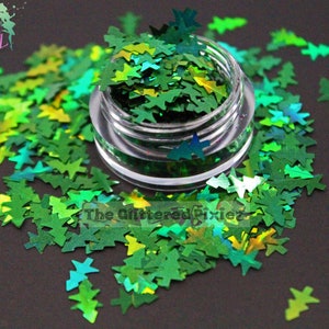 May include: A small jar filled with green and iridescent glitter shaped like Christmas trees. The glitter is scattered around the jar on a black surface.