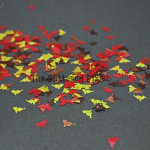 Może przedstawiać: Rozrzucone jesienne liście w odcieniach czerwieni, żółci i brązu. Liście mają kształt małych trójkątów i idealnie nadają się do dodania jesiennego akcentu do rękodzieła i dekoracji.