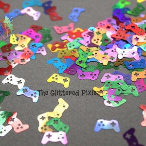 May include: A large assortment of colourful iridescent glitter game controller shapes. The shapes are scattered on a grey surface.