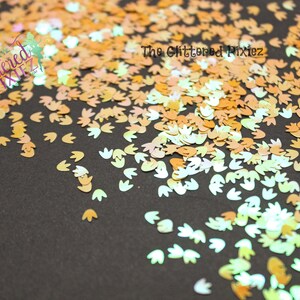 May include: A scattering of iridescent, holographic, and opaque glitter in the shape of a footprint. The glitter is in shades of peach, gold, and white.