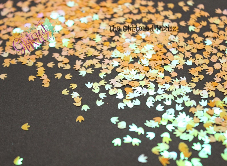 May include: A scattering of iridescent, holographic, and opaque glitter in the shape of a footprint. The glitter is in shades of peach, gold, and white.