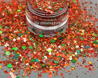 Könnte beinhalten: Nahaufnahme eines Glases mit rotem, orangefarbenem, grünem und goldenem Glitter. Der Glitter ist um das Glas herum auf einer grauen Oberfläche verstreut. Das Glas trägt ein Etikett mit der Aufschrift "The Glittered Pixiez".