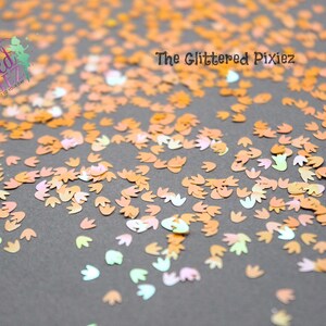 May include: A close-up image of iridescent orange, pink, and green dinosaur footprint shaped glitter. The glitter is scattered on a gray surface. The text "The Glittered Pixiez" is visible in the top center of the image.