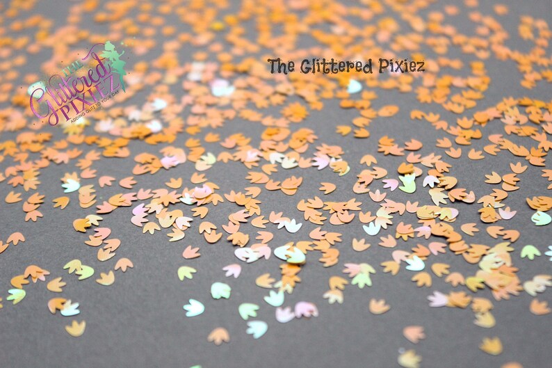 May include: A close-up image of iridescent orange, pink, and green dinosaur footprint shaped glitter. The glitter is scattered on a gray surface. The text "The Glittered Pixiez" is visible in the top center of the image.