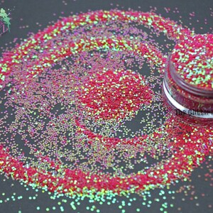 Puede incluir: Primer plano de un tarro de purpurina iridiscente roja y verde. La purpurina está esparcida alrededor del tarro sobre una superficie negra. El tarro tiene una tapa transparente y una etiqueta que dice "The Glittered Pixiez".