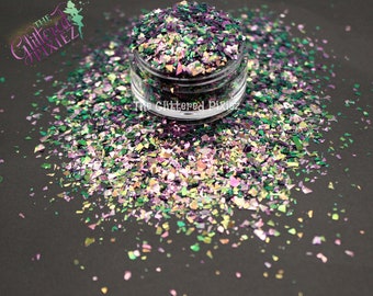May include: A clear jar filled with iridescent glitter flakes in shades of green, purple, and gold. The glitter is scattered around the jar on a dark surface. The text "The Glittered Pixiez" is visible.