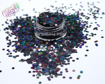 Pode incluir: Um pequeno frasco de glitter iridescente preto, verde, azul e vermelho. O glitter tem a forma de hexágonos. O frasco está etiquetado com "The Glittered Pixiez".