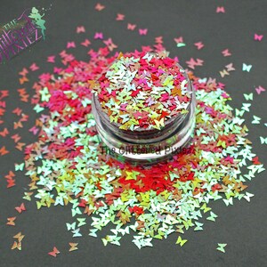 Puede incluir: Un tarro de purpurina de mariposa iridiscente con un arcoíris de colores, incluyendo rojo, rosa, verde y azul. La purpurina está esparcida alrededor del tarro sobre una superficie negra.