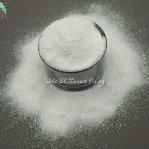 May include: A close-up of a white iridescent glitter in a small glass jar. The glitter is scattered around the jar, creating a sparkling effect.