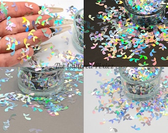 May include: A close-up of iridescent holographic glitter in the shape of leaves. The glitter is loose and scattered on a white surface. The glitter is in a small glass jar.
