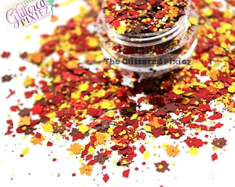 Op de afbeelding: Een potje gevuld met herfstthema glitter in tinten rood, geel en bruin. De glitter is verspreid rond de pot, met blad- en ruitvormen. De tekst "The Glittered Pixiez" is zichtbaar.
