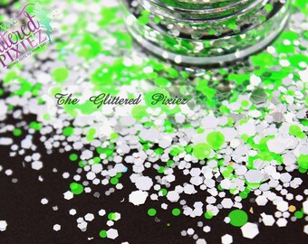 May include: A close-up of green, white, and silver glitter. The glitter is scattered on a black surface. The text "The Glittered Pixiez" is visible in the image.