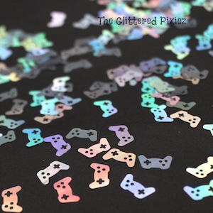 May include: A scattering of iridescent holographic gamer controller confetti in various colors on a black background. The confetti is shaped like a video game controller.