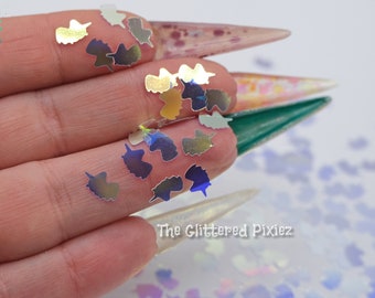 May include: A hand holding iridescent unicorn-shaped glitter. The glitter is scattered on a white surface. The Glitterered Pixiez logo is visible in the bottom right corner.