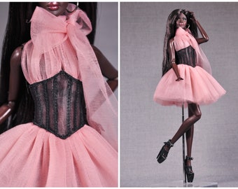 May include: A black doll wearing a pink tulle dress with a black corset and a pink tulle bow around the neck.