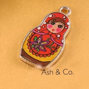 May include: A silver charm with a red, yellow, and white painted design of a Russian nesting doll. The doll has a brown hair and a pink face with black eyes and a red mouth.