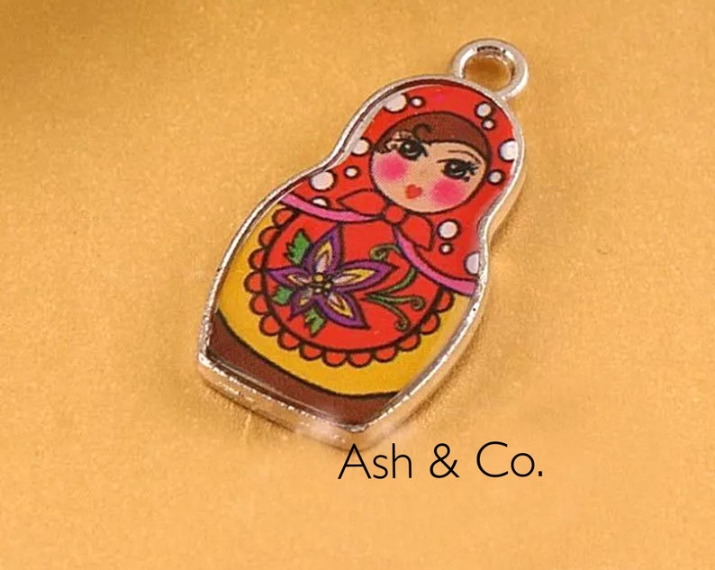 May include: A silver charm with a red, yellow, and white painted design of a Russian nesting doll. The doll has a brown hair and a pink face with black eyes and a red mouth.
