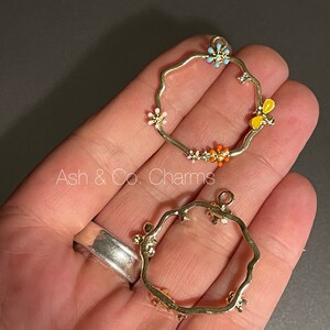 Pode incluir: Dois pingentes de metal dourado com uma forma ondulada irregular. Cada pingente tem um design diferente de flores de esmalte colorido. Os pingentes são para a fabricação de joias.