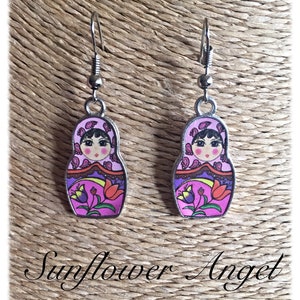 May include: A pair of silver dangle earrings featuring a pink and purple Russian nesting doll design. The dolls have black hair, brown eyes, and a floral pattern on their dresses.