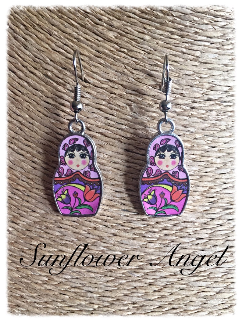 May include: A pair of silver dangle earrings featuring a pink and purple Russian nesting doll design. The dolls have black hair, brown eyes, and a floral pattern on their dresses.