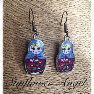 May include: A pair of silver dangle earrings featuring a Russian nesting doll design. The dolls are painted with a blue, white, and red floral pattern. The earrings are a whimsical and colorful accessory.