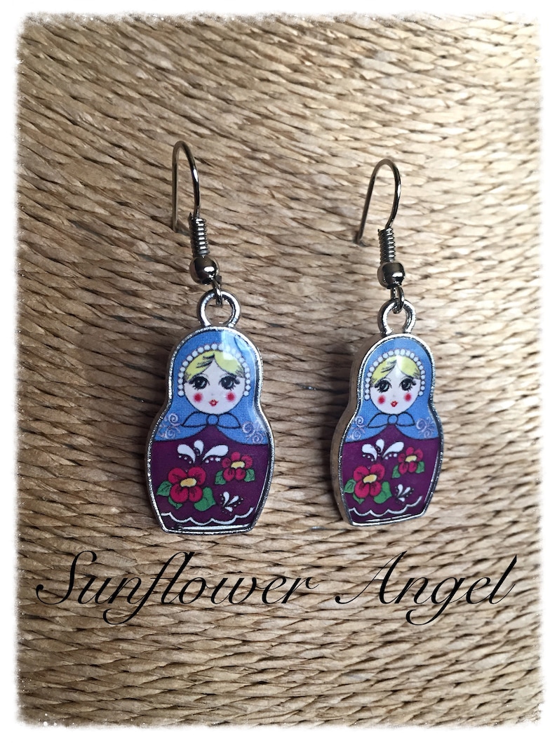 May include: A pair of silver dangle earrings featuring a Russian nesting doll design. The dolls are painted with a blue, white, and red floral pattern. The earrings are a whimsical and colorful accessory.