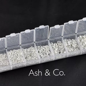 Op de afbeelding: Een doorzichtige plastic organizer gevuld met verschillende maten zilveren sprongringen. De organizer heeft een scharnierend deksel en is verdeeld in verschillende compartimenten. De tekst "Ash & Co." staat onderaan.
