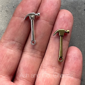 Può includere: Due ciondoli a forma di martello, uno argentato e uno in bronzo. I ciondoli sono piccoli e hanno un anello in basso per attaccarli a una collana o un braccialetto.