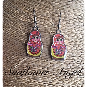 May include: A pair of silver dangle earrings featuring a red, yellow, and white Russian nesting doll design. The dolls have black eyes, pink cheeks, and a purple flower on their dress.