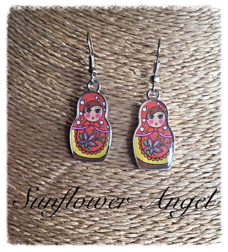 May include: A pair of silver dangle earrings featuring a red, yellow, and white Russian nesting doll design. The dolls have black eyes, pink cheeks, and a purple flower on their dress.