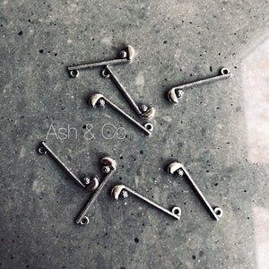 May include: Silver golf club charms on a grey surface. Each charm has a detailed club head and a textured handle, with small loops for attachment. The text "Ash & Co." is visible. Ideal for jewellery making or crafts.
