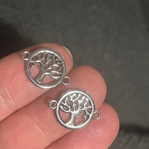 Könnte beinhalten: Zwei silberfarbene Metall-Anhänger in Form eines Lebensbaums. Jeder Anhänger hat ein kreisförmiges Design mit einer detaillierten Baum-Silhouette und zwei Sprungringe zum Anbringen an Schmuck oder anderen Bastelarbeiten.